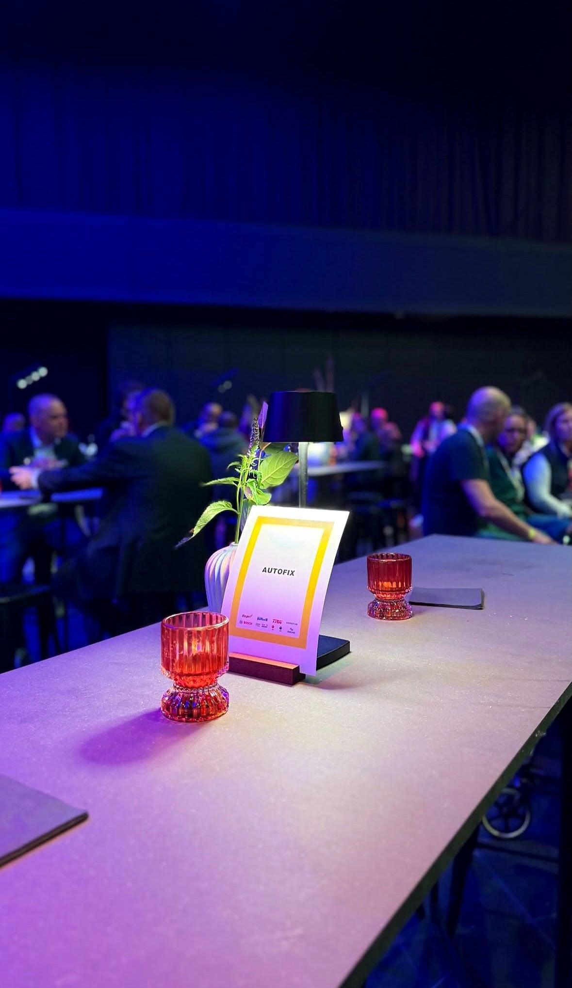 Tafel met een bordje van AUTOFIX en kaarsjes op het event van Groep Doyen op het autosalon, met bezoekers op de achtergrond