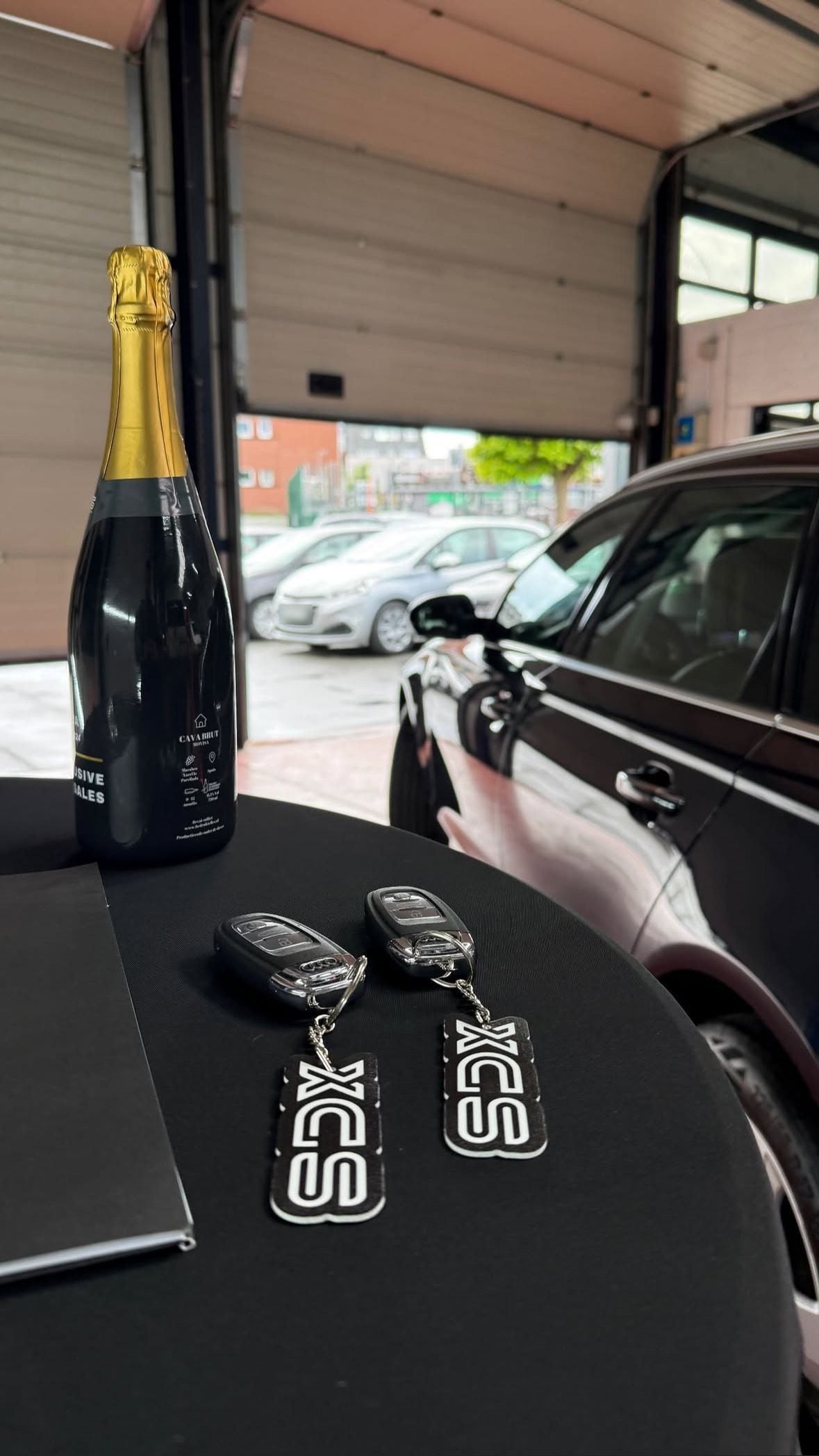 Overdracht van een wagen bij XCS Automotive met champagne en XCS-sleutelhangers